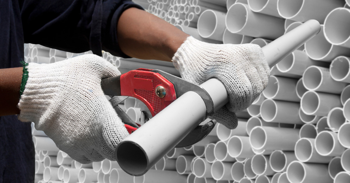 A worker wearing white gloves is using a red cutting tool on a PVC pipe. Many other pipes are stacked nearby.