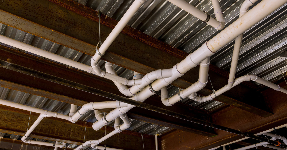 A large, white PVC plumbing system weaves across a metal ceiling with support beams in a dark room.