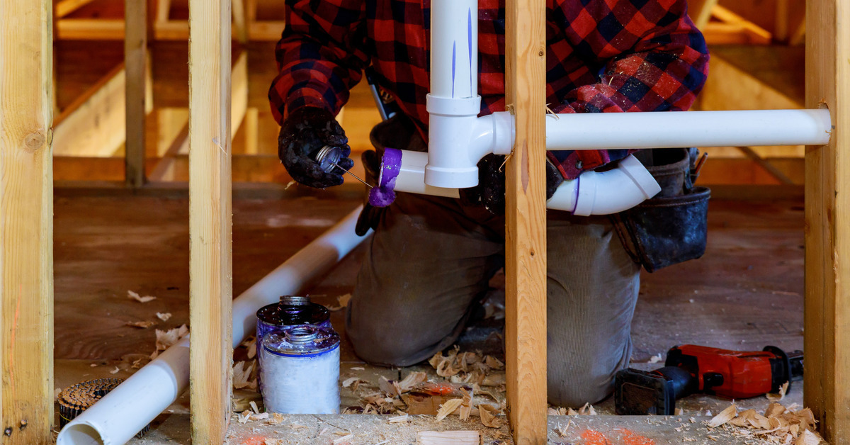 A worker is applying purple sealant to a gray PVC pipe fitting that is inside of a construction site.