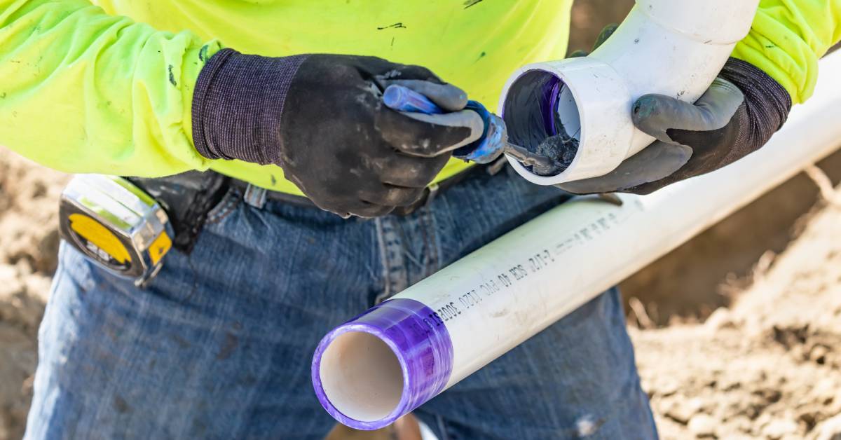 A person wearing a neon green shirt and black gloves is applying PVC cement at the edge of an elbow pipe fitting.