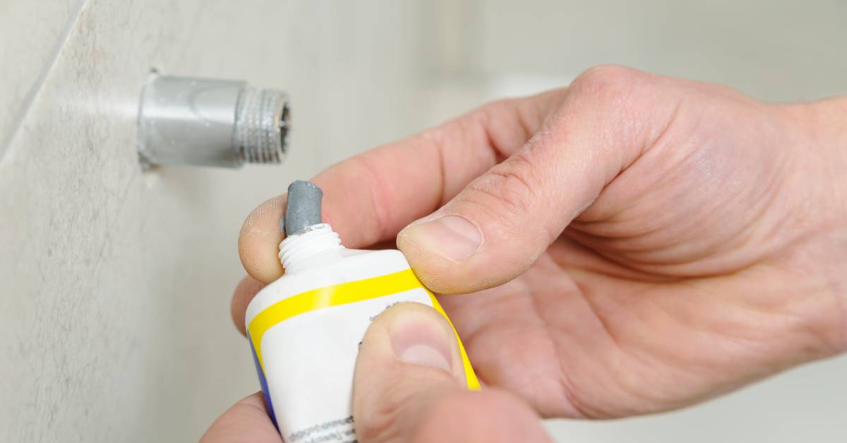 A person is squeezing a gray sealant out of a small tube. The person is standing in front of a metal pipe.