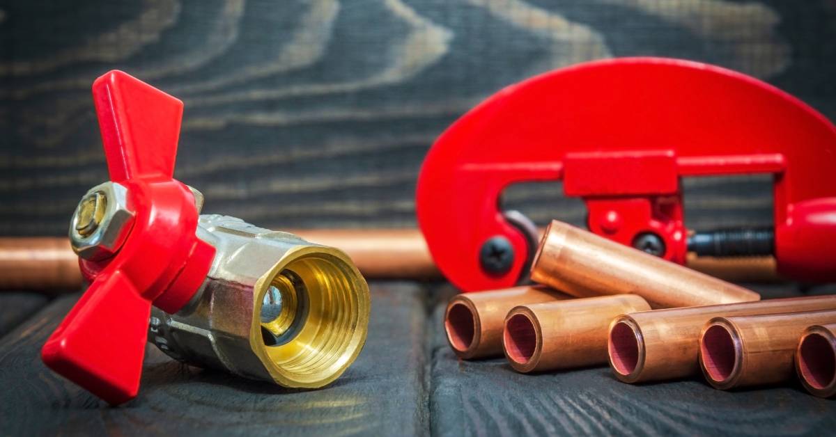 A compact ball valve with a red handle sitting on a wooden surface next to six brass pipes and a large red component.