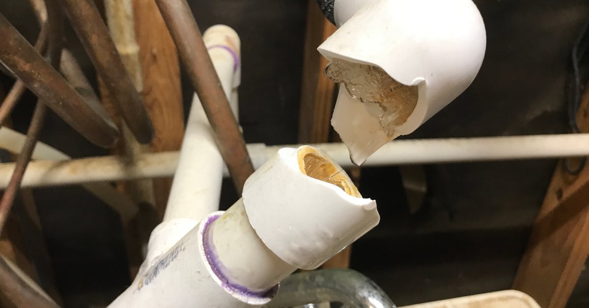 A burst white PVC pipe installed in a cluttered space. Ice is sticking out of the cracked ends of the PVC.