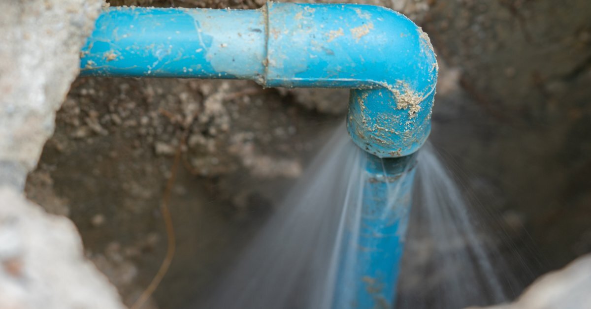  A blue pipe system is covered in dirt and sticking out of a hole. Water is bursting from one end of a pipe joint.