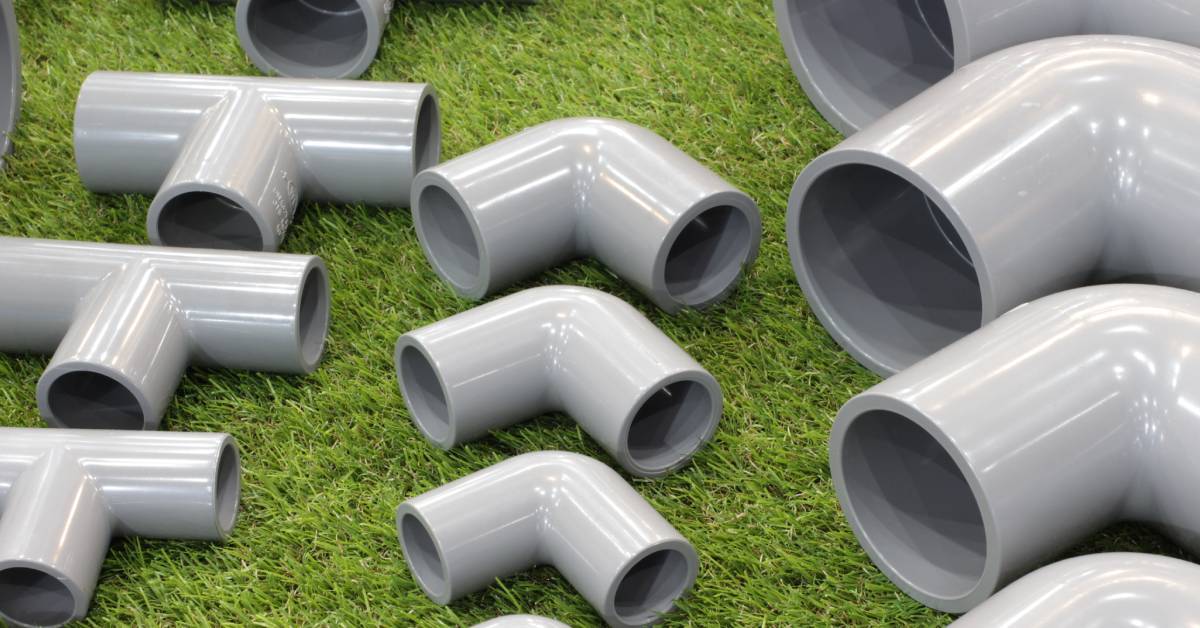 A large variety of gray PVC pipe fittings are neatly spread apart outside on a bright green patch of grass.