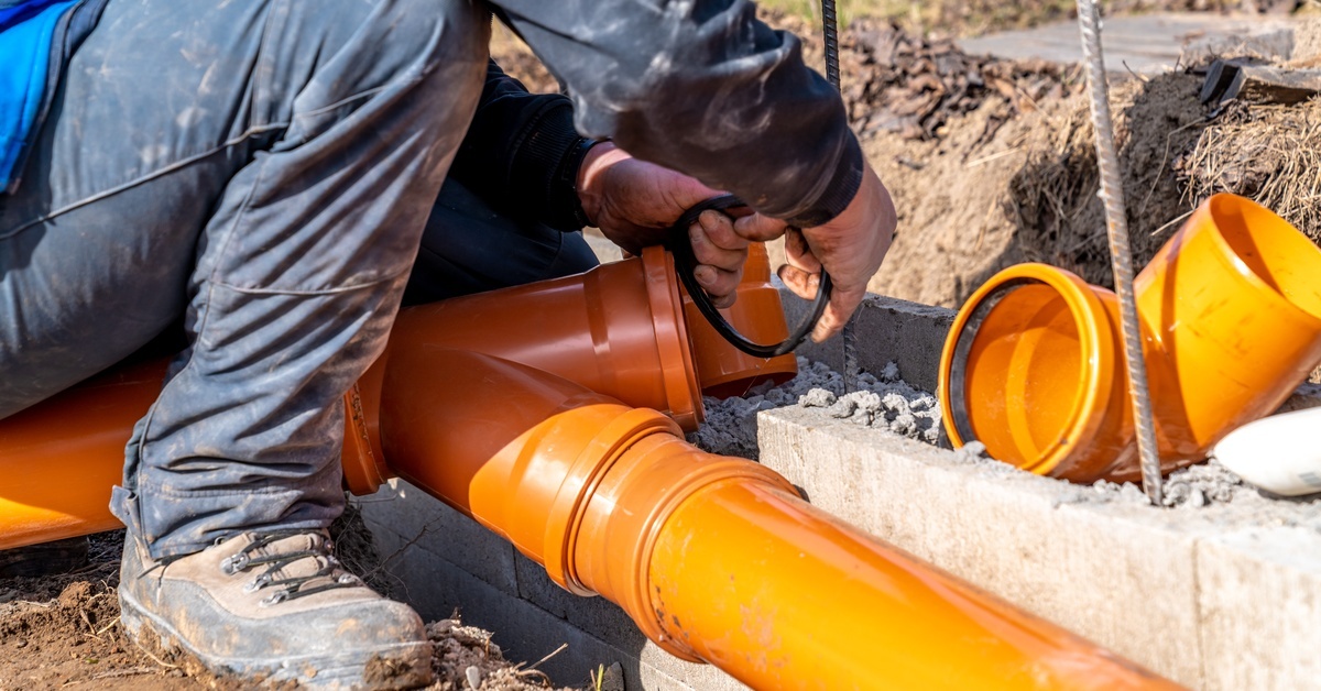 A person wearing boots is crouching down next to orange PVC pipes and disassembling them during the day.